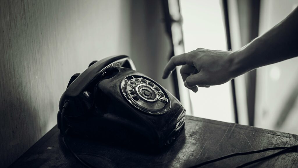A monochrome photo of a hand reaching towards a vintage black rotary telephone in an indoor setting.
