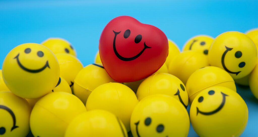 A red heart-shaped smiling ball stands out among yellow smiley faces, symbolizing individuality.